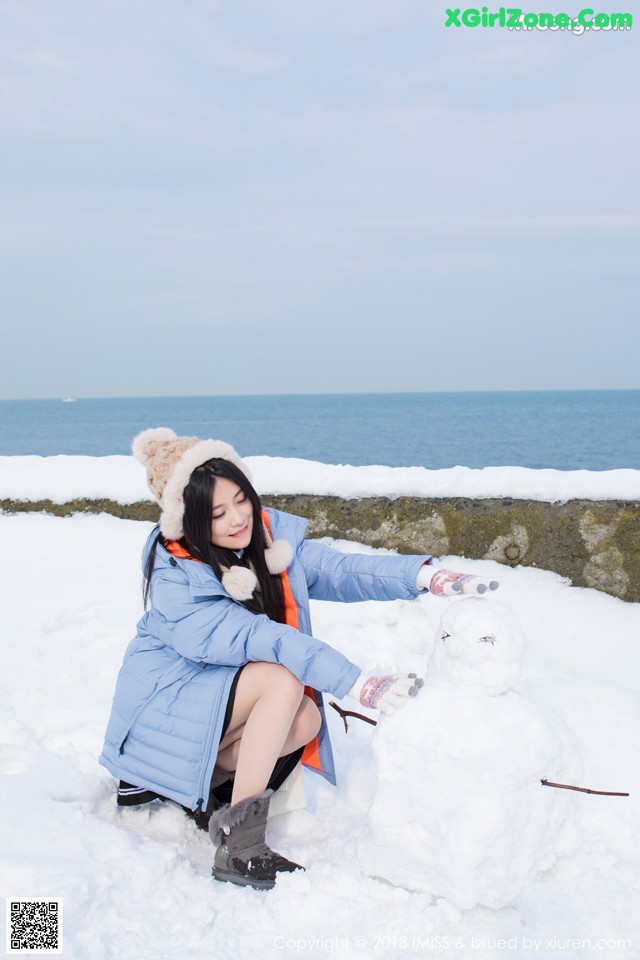 A woman kneeling down next to a snowman in the snow.