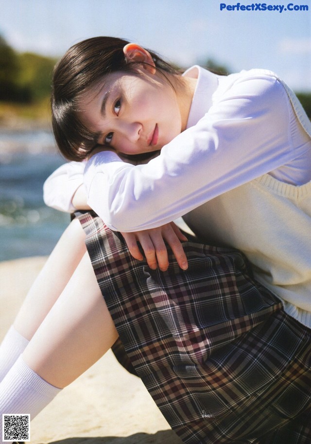 A woman in a school uniform sitting on a rock by the water.